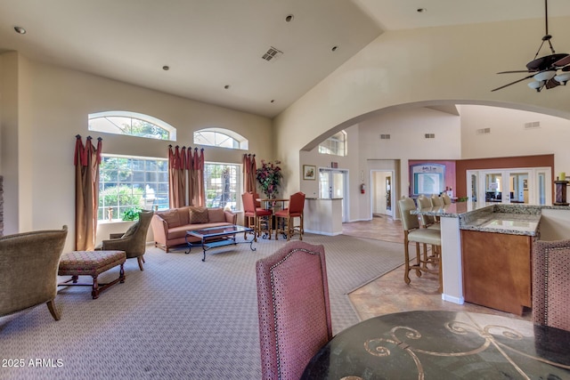 living room featuring a ceiling fan, visible vents, high vaulted ceiling, arched walkways, and light carpet