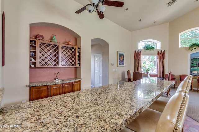 kitchen featuring visible vents, ceiling fan, light stone countertops, brown cabinets, and arched walkways