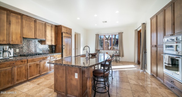 kitchen with appliances with stainless steel finishes, tasteful backsplash, sink, dark stone countertops, and a center island with sink