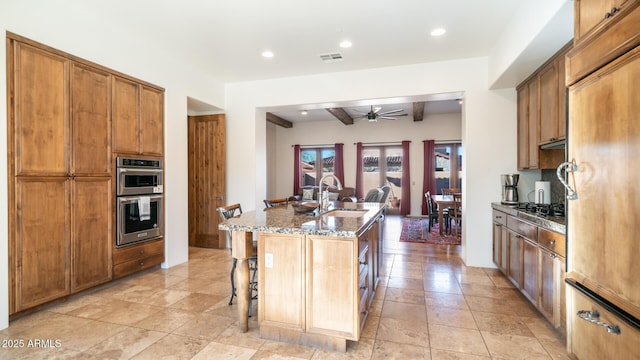 kitchen featuring beamed ceiling, a kitchen bar, sink, a center island with sink, and double oven