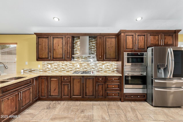 kitchen with wall chimney range hood, decorative backsplash, appliances with stainless steel finishes, a sink, and stone tile flooring
