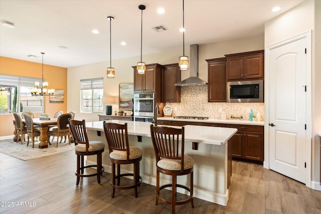 kitchen featuring appliances with stainless steel finishes, pendant lighting, an island with sink, a kitchen breakfast bar, and wall chimney range hood