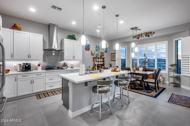 kitchen featuring white cabinets, decorative light fixtures, a center island with sink, and wall chimney exhaust hood