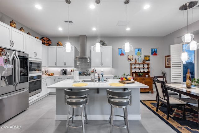 kitchen featuring a kitchen island with sink, white cabinets, hanging light fixtures, wall chimney exhaust hood, and stainless steel appliances