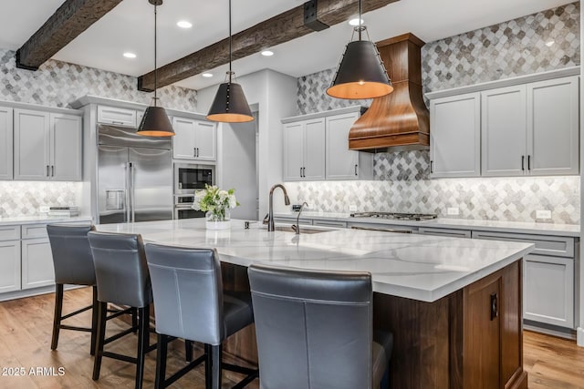 kitchen featuring sink, hanging light fixtures, built in appliances, an island with sink, and custom range hood