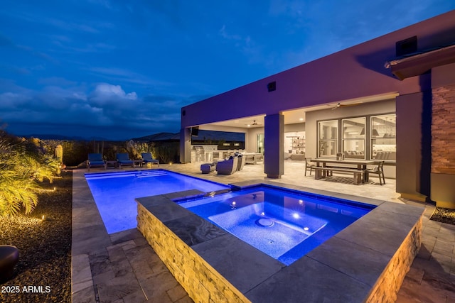 pool at dusk with an in ground hot tub, an outdoor kitchen, ceiling fan, and a patio area