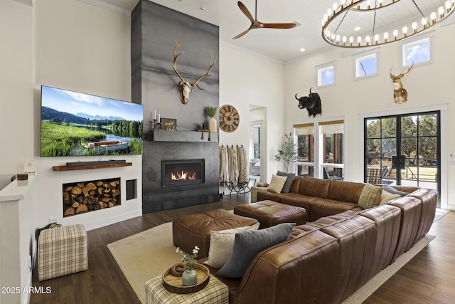 living area with a warm lit fireplace, wood-type flooring, ceiling fan, ornamental molding, and a high ceiling