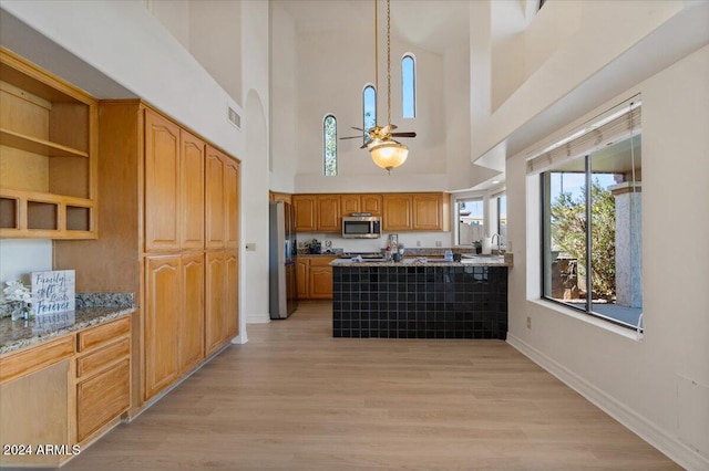 kitchen featuring light wood-type flooring, light stone counters, stainless steel appliances, ceiling fan, and a high ceiling