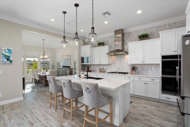 kitchen with white cabinetry, hanging light fixtures, an island with sink, stainless steel appliances, and wall chimney exhaust hood