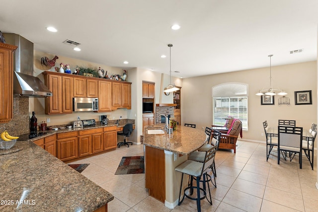 kitchen with light tile patterned floors, visible vents, brown cabinets, wall chimney exhaust hood, and stainless steel microwave