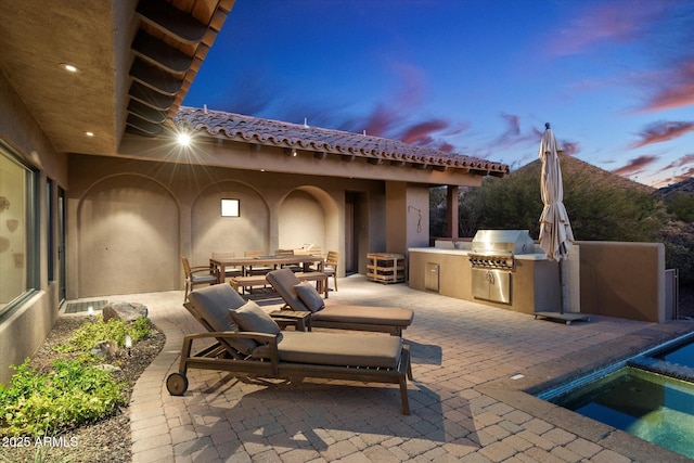 patio terrace at dusk with an in ground hot tub, area for grilling, and exterior kitchen