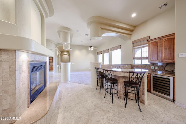 kitchen featuring a breakfast bar, beverage cooler, backsplash, a tiled fireplace, and ceiling fan