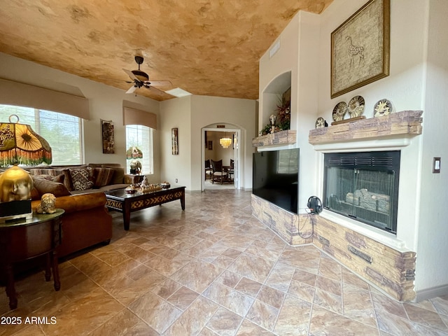 living area featuring ceiling fan, arched walkways, a glass covered fireplace, and baseboards