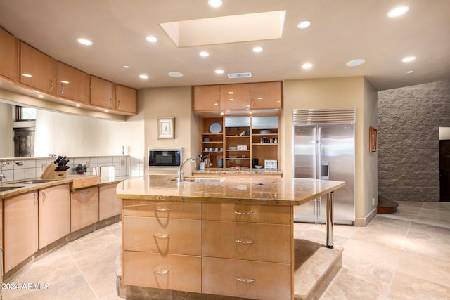 kitchen with an island with sink, built in appliances, sink, a skylight, and stone countertops