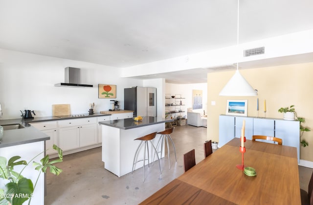 kitchen featuring white cabinets, stainless steel fridge, a kitchen bar, hanging light fixtures, and wall chimney range hood