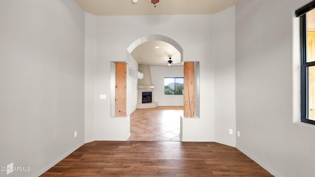 spare room with a fireplace with raised hearth, a ceiling fan, wood finished floors, arched walkways, and baseboards