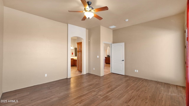 unfurnished bedroom featuring ensuite bath, wood finished floors, arched walkways, baseboards, and ceiling fan