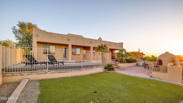 back of property at dusk with fence, a yard, a fireplace, stucco siding, and a patio area