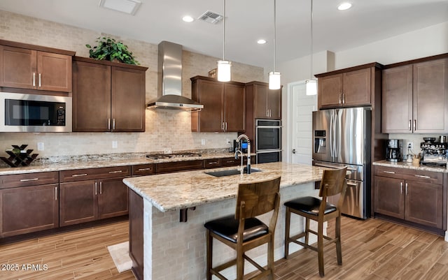 kitchen with light stone counters, a center island with sink, pendant lighting, stainless steel appliances, and wall chimney range hood