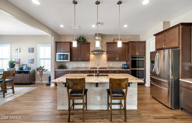 kitchen featuring wall chimney exhaust hood, appliances with stainless steel finishes, an island with sink, pendant lighting, and light stone countertops