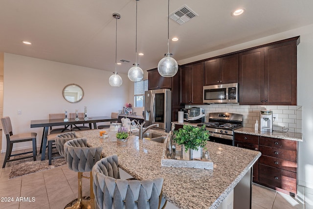 kitchen with hanging light fixtures, light stone countertops, an island with sink, appliances with stainless steel finishes, and light tile patterned flooring