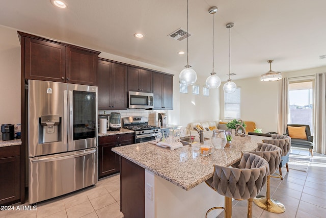 kitchen with appliances with stainless steel finishes, backsplash, light stone counters, a center island with sink, and decorative light fixtures