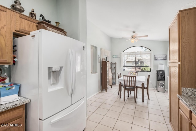 kitchen with white refrigerator with ice dispenser, a ceiling fan, light tile patterned floors, stone countertops, and brown cabinetry