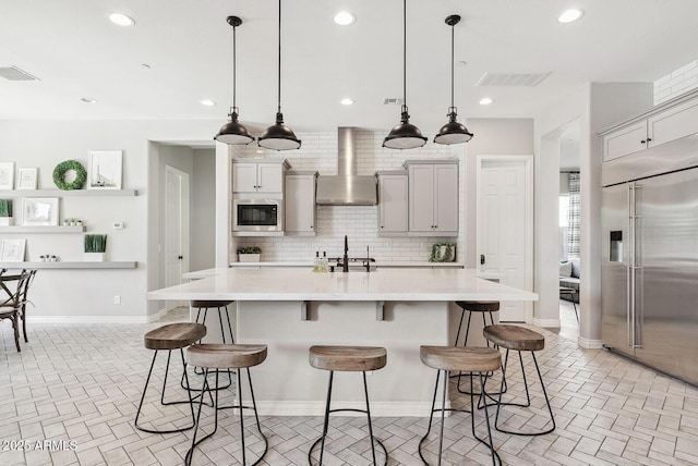 kitchen featuring a breakfast bar, built in appliances, light countertops, wall chimney exhaust hood, and tasteful backsplash