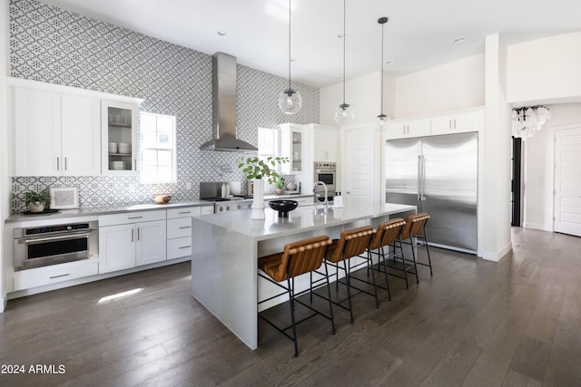 kitchen with white cabinetry, stainless steel appliances, wall chimney range hood, decorative light fixtures, and a kitchen island with sink