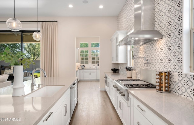 kitchen with stainless steel appliances, sink, wall chimney range hood, pendant lighting, and white cabinetry