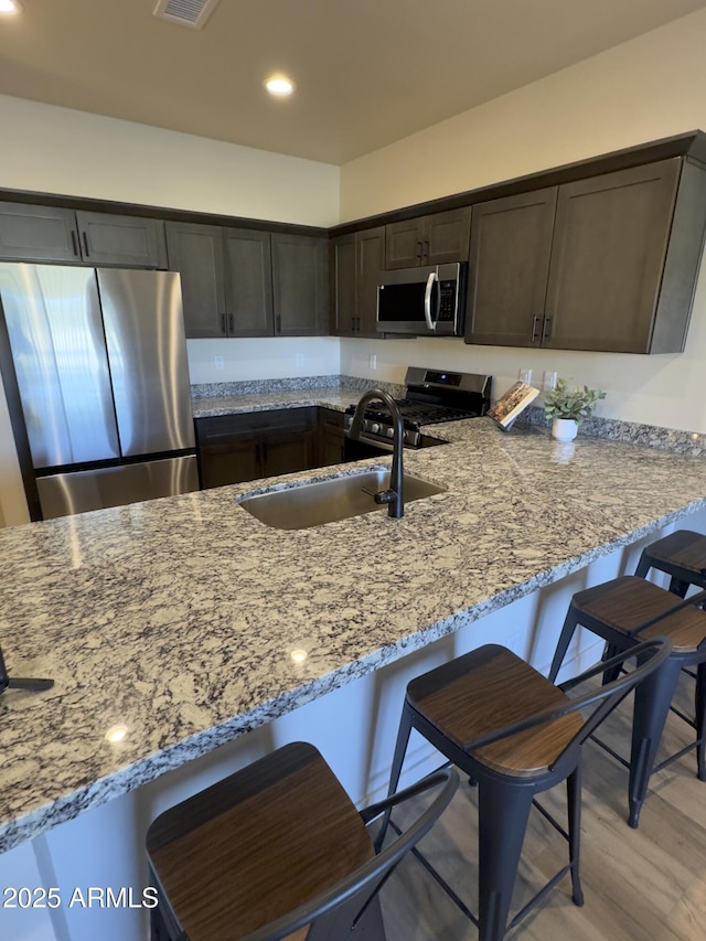 kitchen with light stone counters, stainless steel appliances, dark brown cabinetry, a sink, and a peninsula