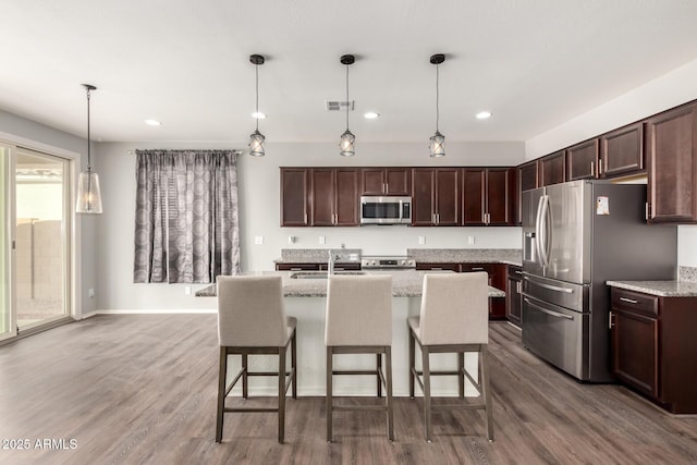 kitchen with appliances with stainless steel finishes, hardwood / wood-style floors, pendant lighting, an island with sink, and light stone countertops