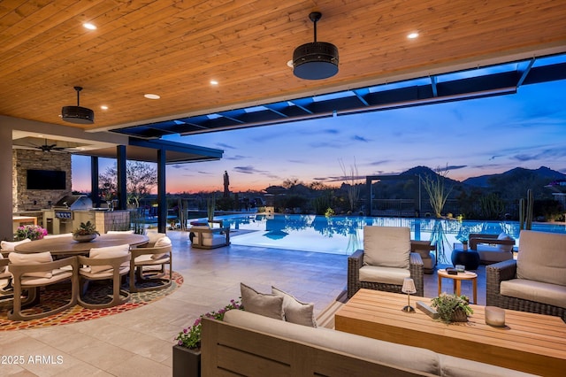 8328 E Whisper Rock Trl, Scottsdale, AZ 85266 - pool at dusk with an outdoor hangout area, an outdoor pool, exterior kitchen, a patio area, and a ceiling fan