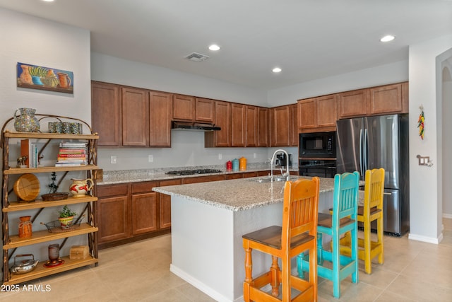 kitchen with sink, a breakfast bar area, black appliances, light stone countertops, and a center island with sink