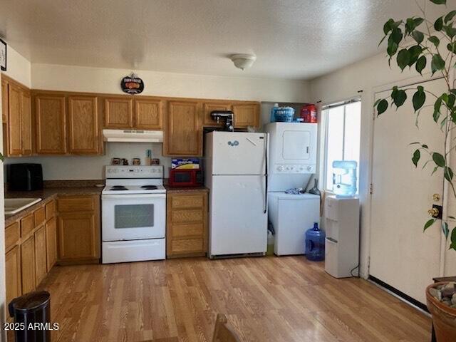 kitchen with light wood finished floors, brown cabinets, stacked washing maching and dryer, under cabinet range hood, and white appliances