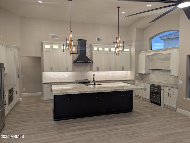 kitchen featuring wall chimney range hood, beverage cooler, a sink, tasteful backsplash, and a center island with sink