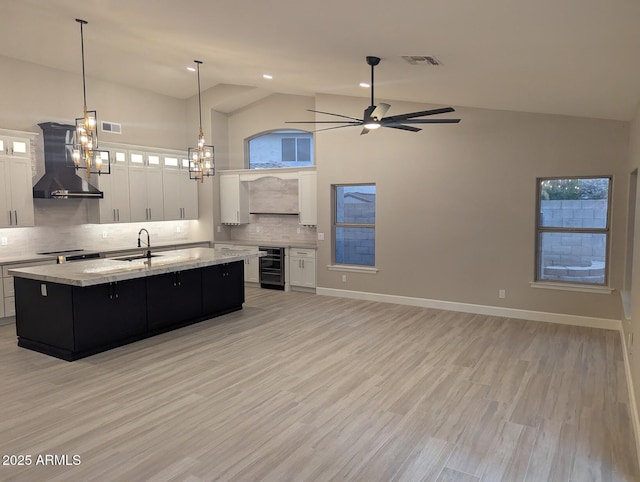 kitchen with wall chimney exhaust hood, decorative backsplash, a center island with sink, a ceiling fan, and white cabinets