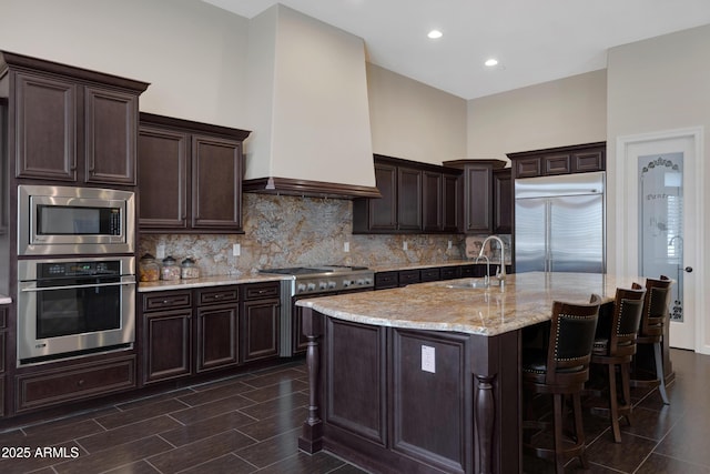 kitchen with dark brown cabinetry, light stone counters, built in appliances, a kitchen island with sink, and a sink