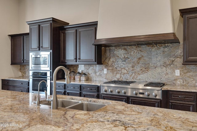 kitchen with dark brown cabinetry, stainless steel appliances, a sink, decorative backsplash, and custom range hood