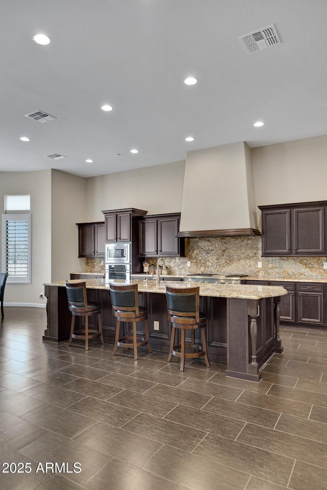 kitchen featuring dark brown cabinetry, visible vents, custom range hood, a kitchen island with sink, and stainless steel appliances