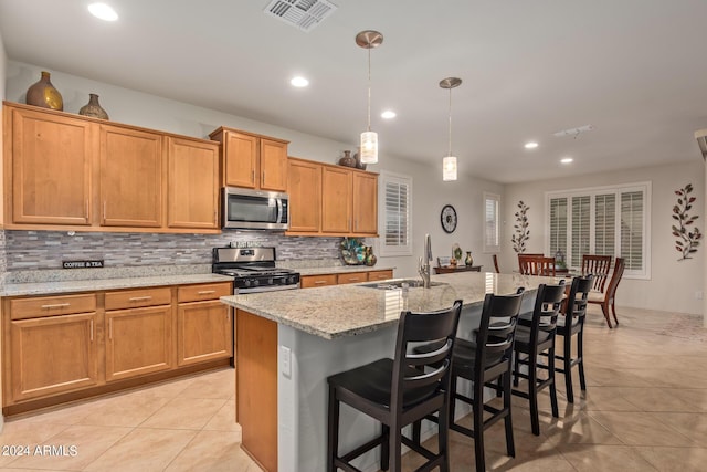 kitchen featuring sink, appliances with stainless steel finishes, a kitchen island with sink, hanging light fixtures, and light stone countertops