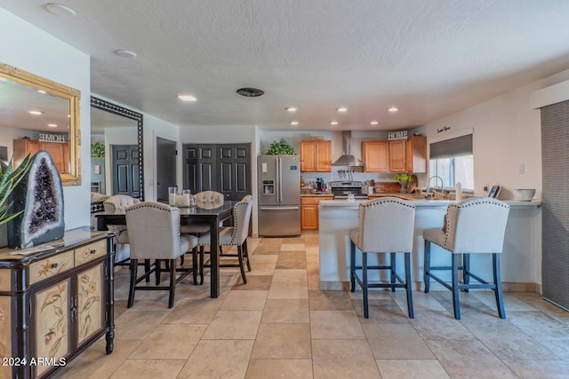 kitchen with wall chimney exhaust hood, a breakfast bar area, a textured ceiling, kitchen peninsula, and stainless steel appliances