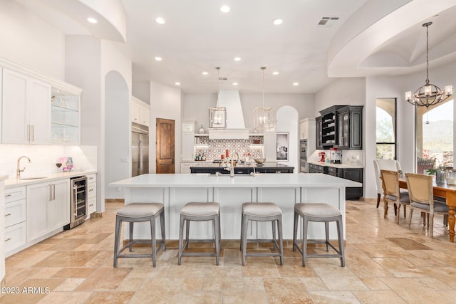 kitchen featuring custom range hood, beverage cooler, a large island with sink, decorative light fixtures, and white cabinetry