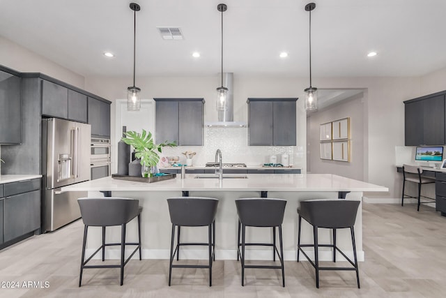 kitchen featuring pendant lighting, stainless steel appliances, wall chimney exhaust hood, and an island with sink