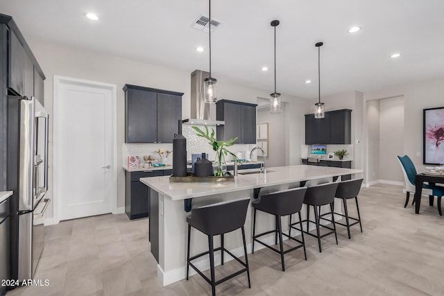 kitchen featuring a center island with sink, backsplash, pendant lighting, high quality fridge, and wall chimney exhaust hood