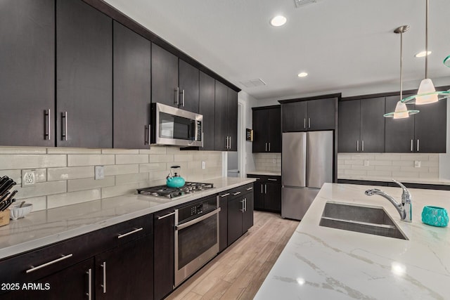 kitchen with appliances with stainless steel finishes, light stone counters, decorative light fixtures, light wood-type flooring, and a sink