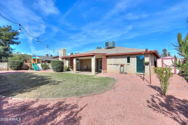 rear view of property with a yard, a patio area, central air condition unit, a playground, and a storage unit