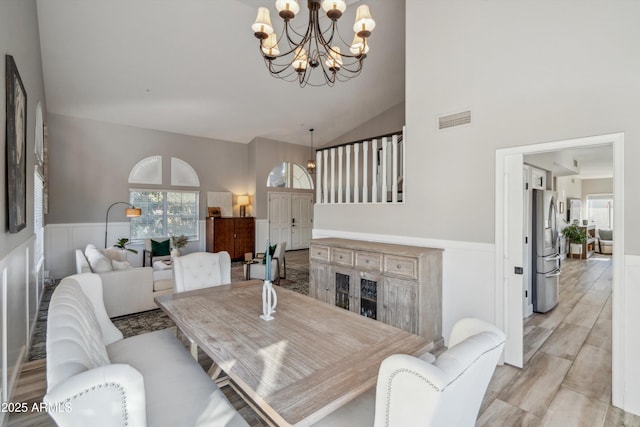 dining space with high vaulted ceiling, light wood-style flooring, a notable chandelier, visible vents, and wainscoting