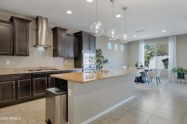 kitchen with wall chimney exhaust hood, dark brown cabinets, hanging light fixtures, an island with sink, and stainless steel appliances