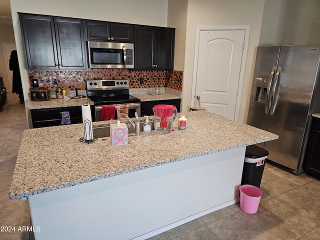 kitchen with light stone countertops, tasteful backsplash, a kitchen island with sink, light tile patterned floors, and appliances with stainless steel finishes